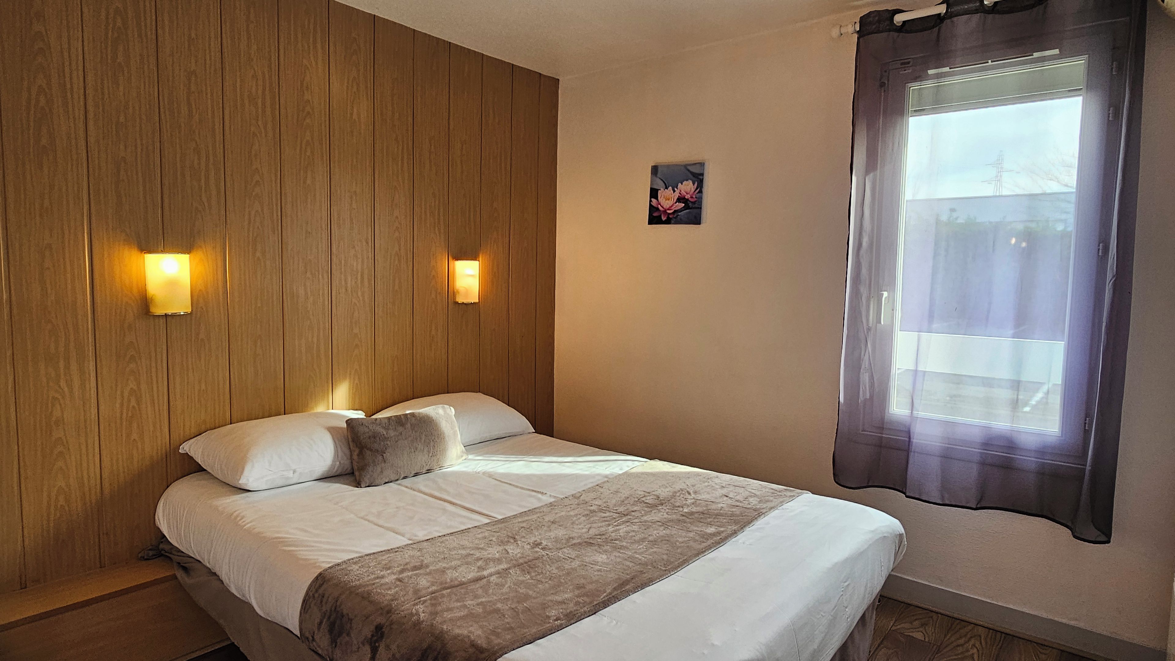 A single bed with a wooden headboard, two wall-mounted lights, a window with a curtain, and a small floral picture on the wall.