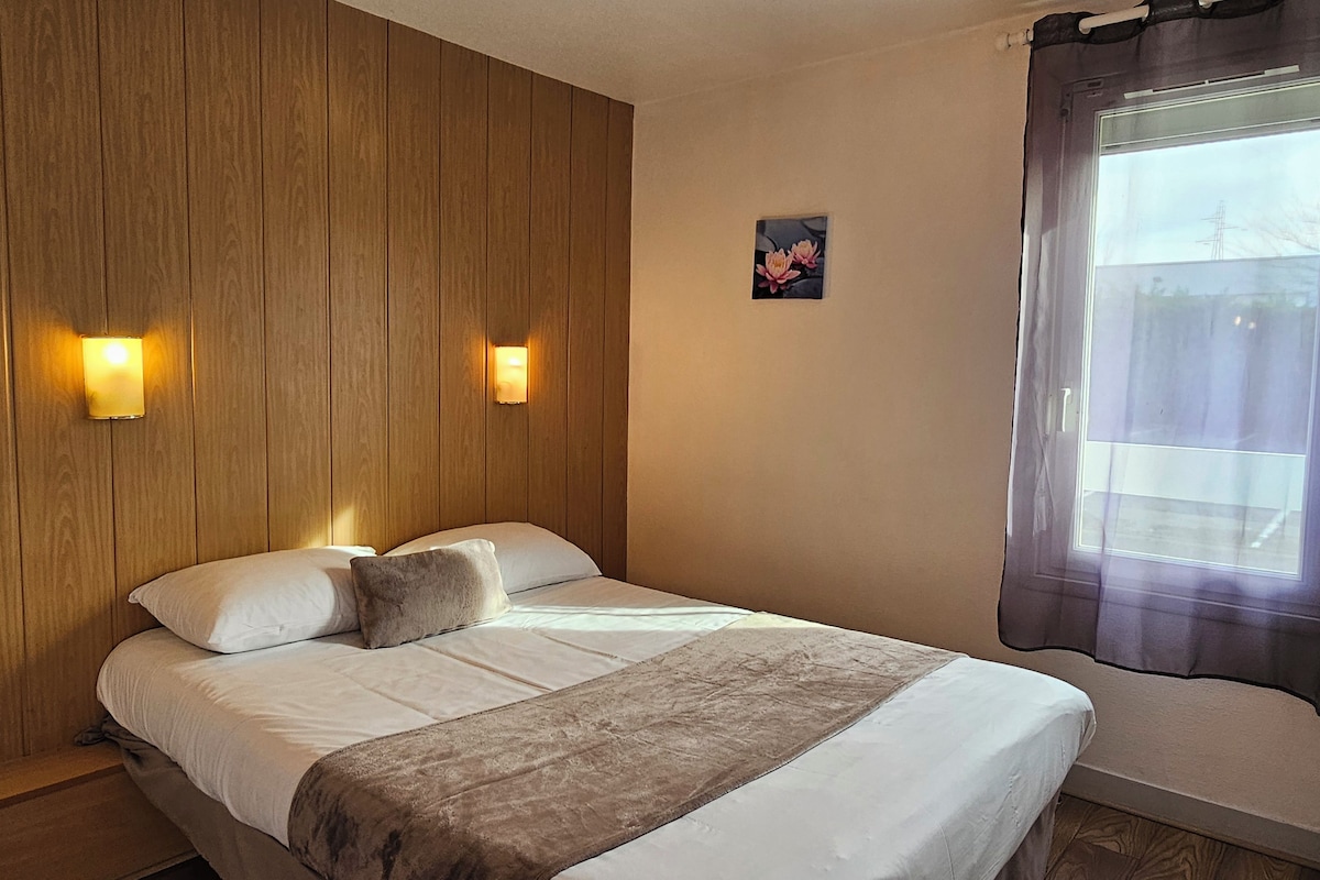 Una cama individual con cabecera de madera, dos lámparas fijadas a la pared, una ventana con cortina y un pequeño cuadro floral.