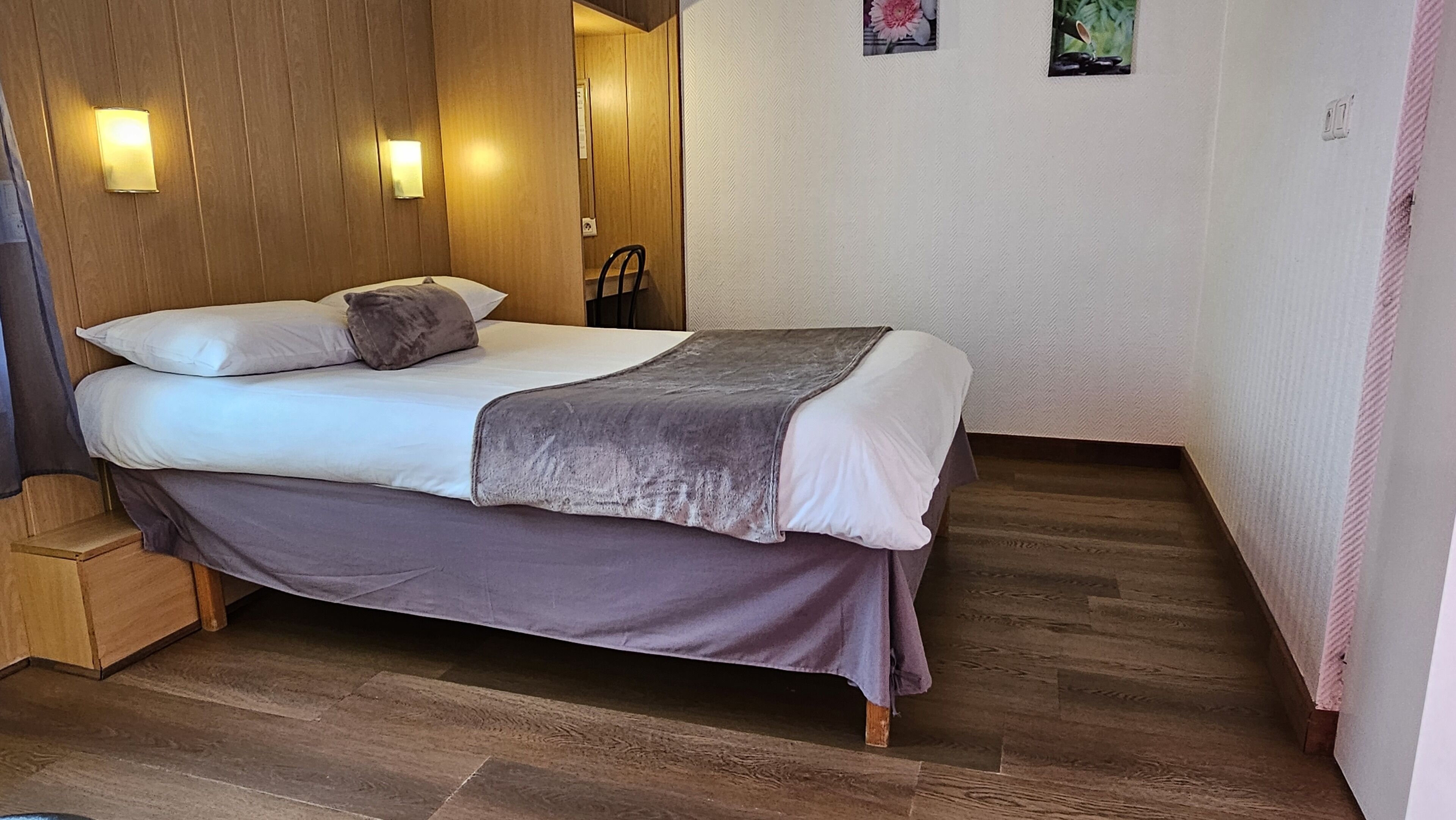 Een net opgemaakt bed met een grijze deken en kussens, houten vloer en een wand met twee ingelijste foto's.