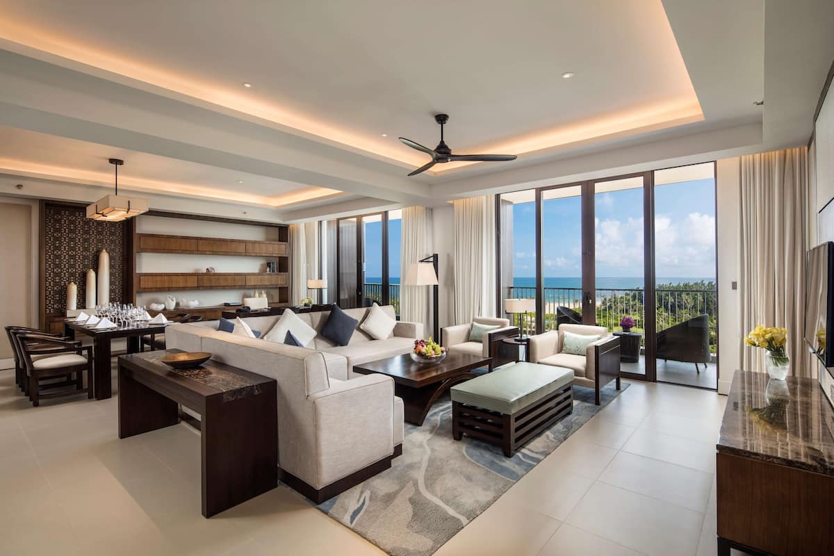 A spacious living room with a large sofa, coffee table, and dining area. It features a ceiling fan, large windows with a view of the ocean, and a flat-screen TV on the wall.
