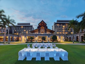 Experience the hotel's unique architecture and tranquil surroundings, featuring a central building with a striking roof structure, surrounded by lush lawns and palm trees.