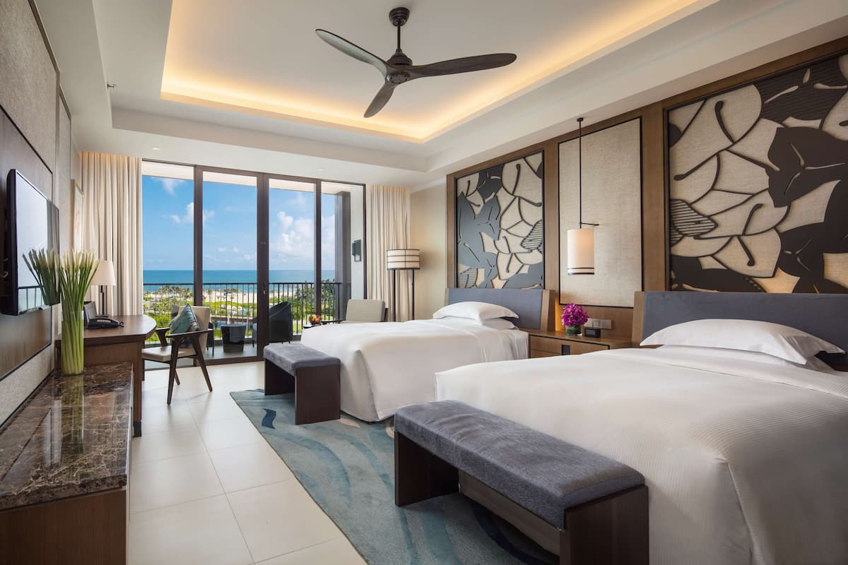 A hotel room with two beds, a desk, a chair, a TV, and a balcony with a view of the ocean.