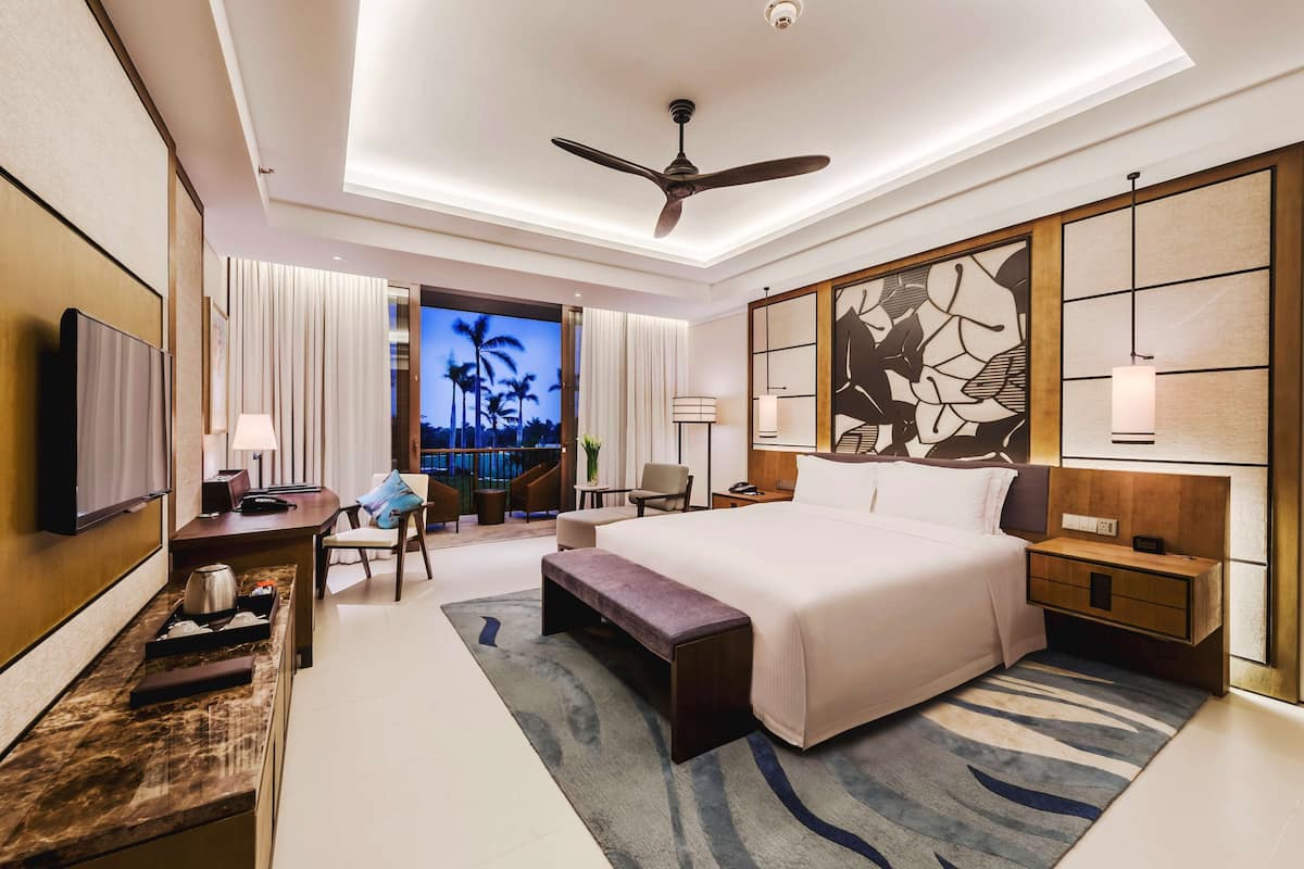 A modern hotel room with a large bed, a desk, a chair, a TV, and a balcony with a view of palm trees.