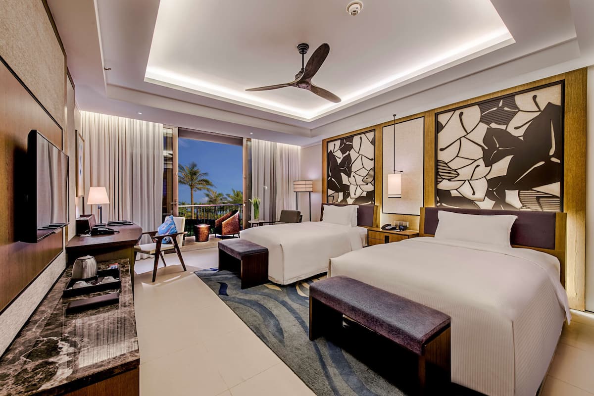A hotel room with two beds, a desk, a chair, a TV, and a balcony with a view of palm trees.