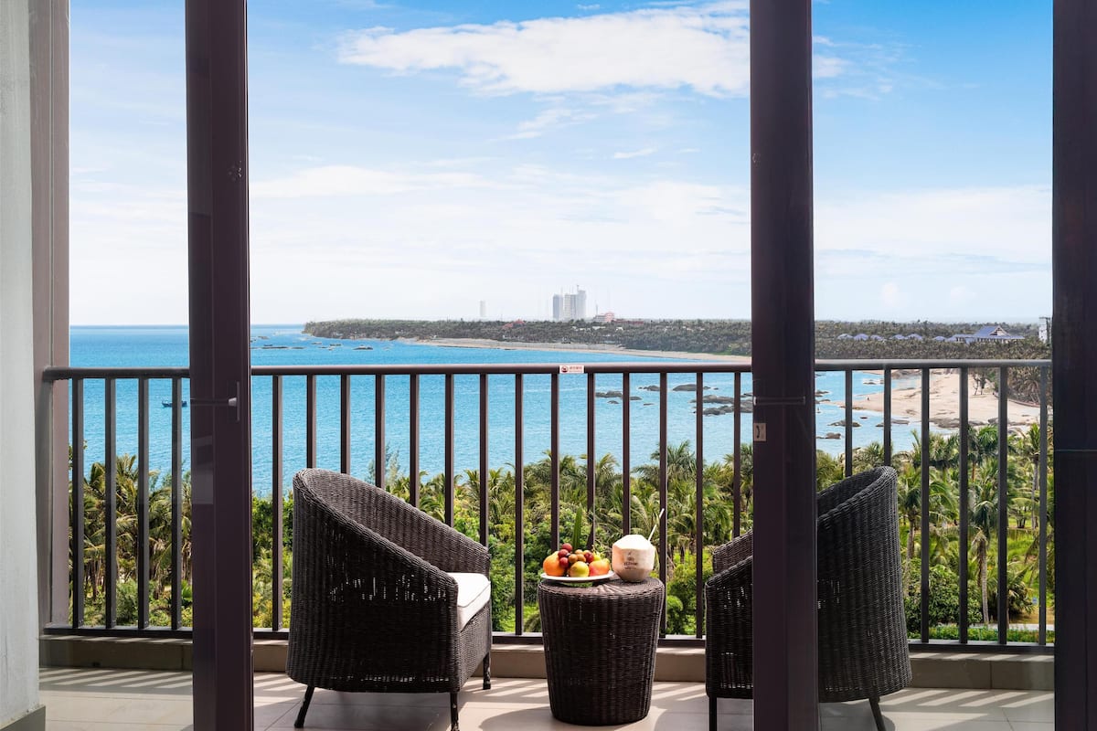 A balcony with wicker chairs, a small table with fruits and a coconut, and a view of the ocean and beach.
