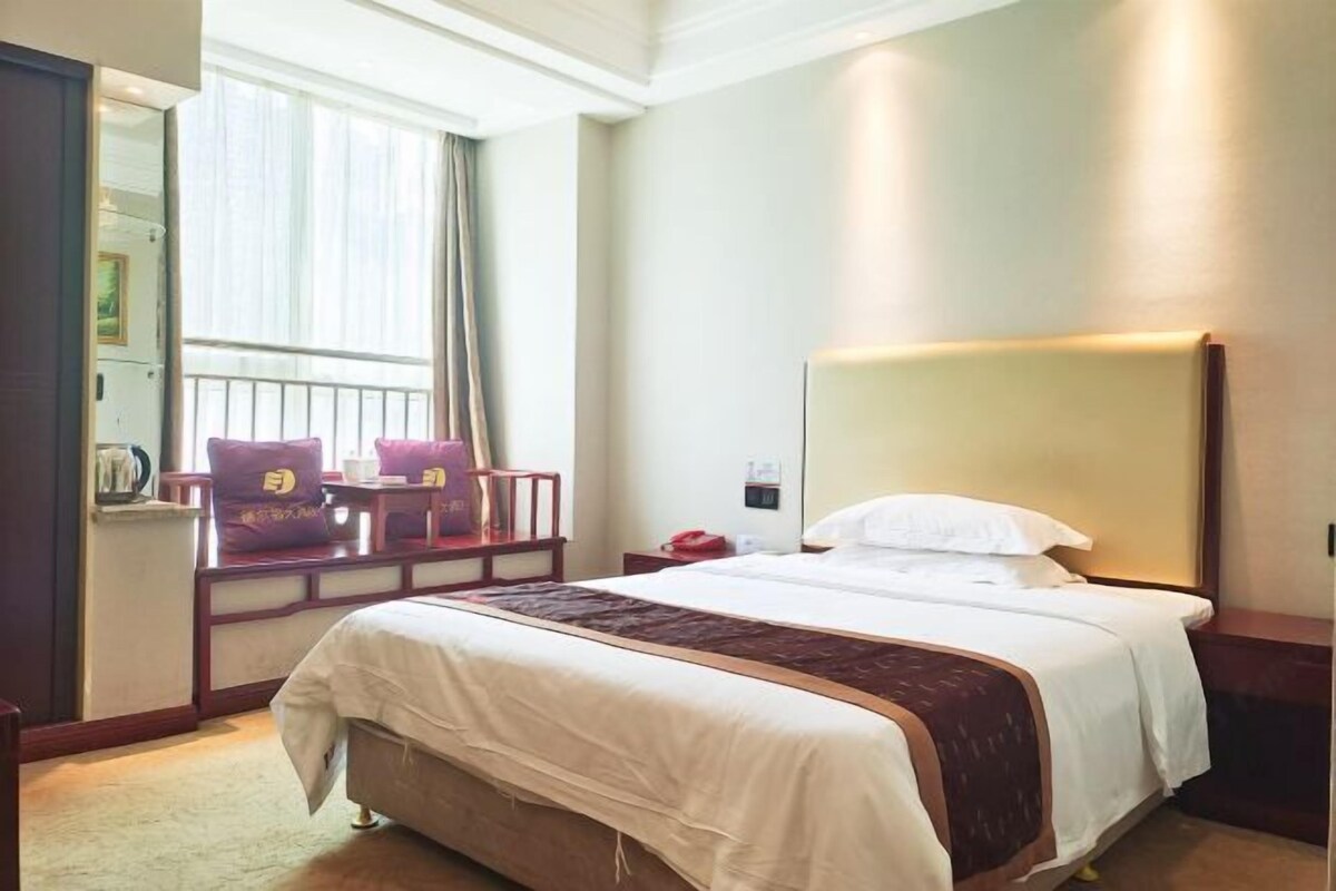A hotel room with a large bed, a bedside table, a chair, a window with curtains, and a wall-mounted lamp.