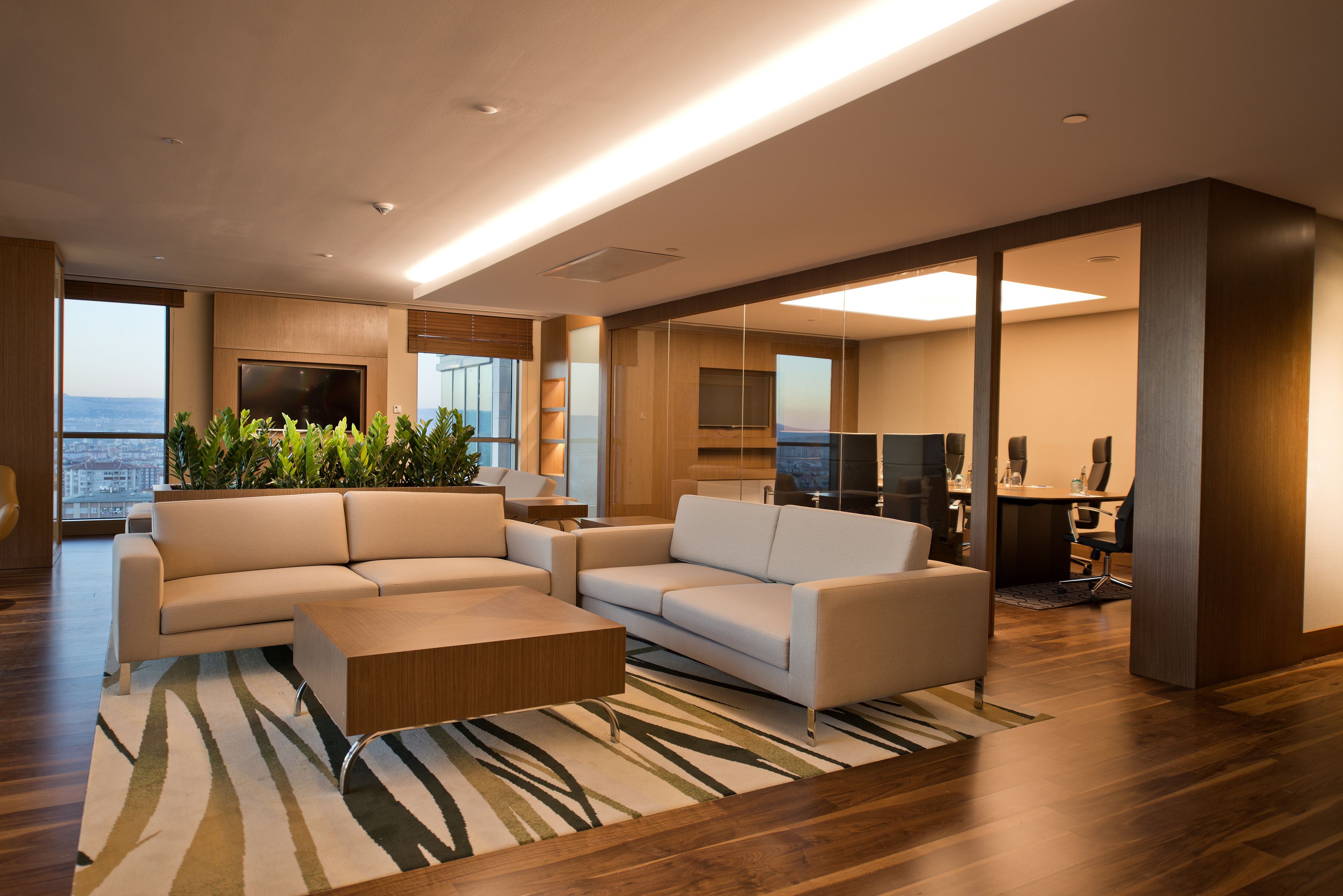 Una sala moderna con un sofá grande, una mesa de centro y una televisión. Se ve la ciudad a través de amplios ventanales.