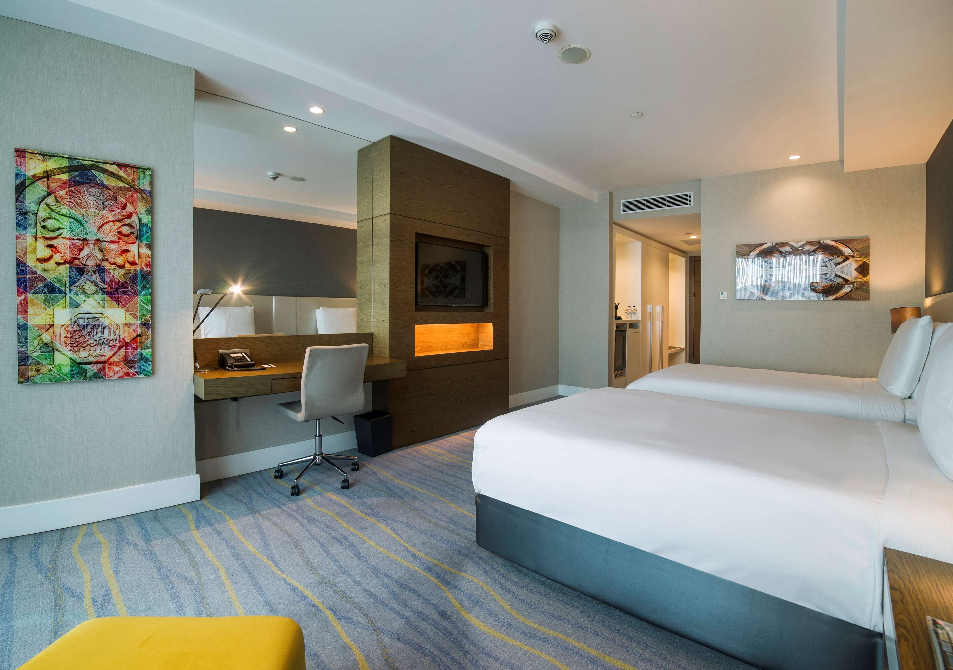 Habitación de hotel con una cama grande, un escritorio con silla, una mesita y una obra de arte colorida en la pared.