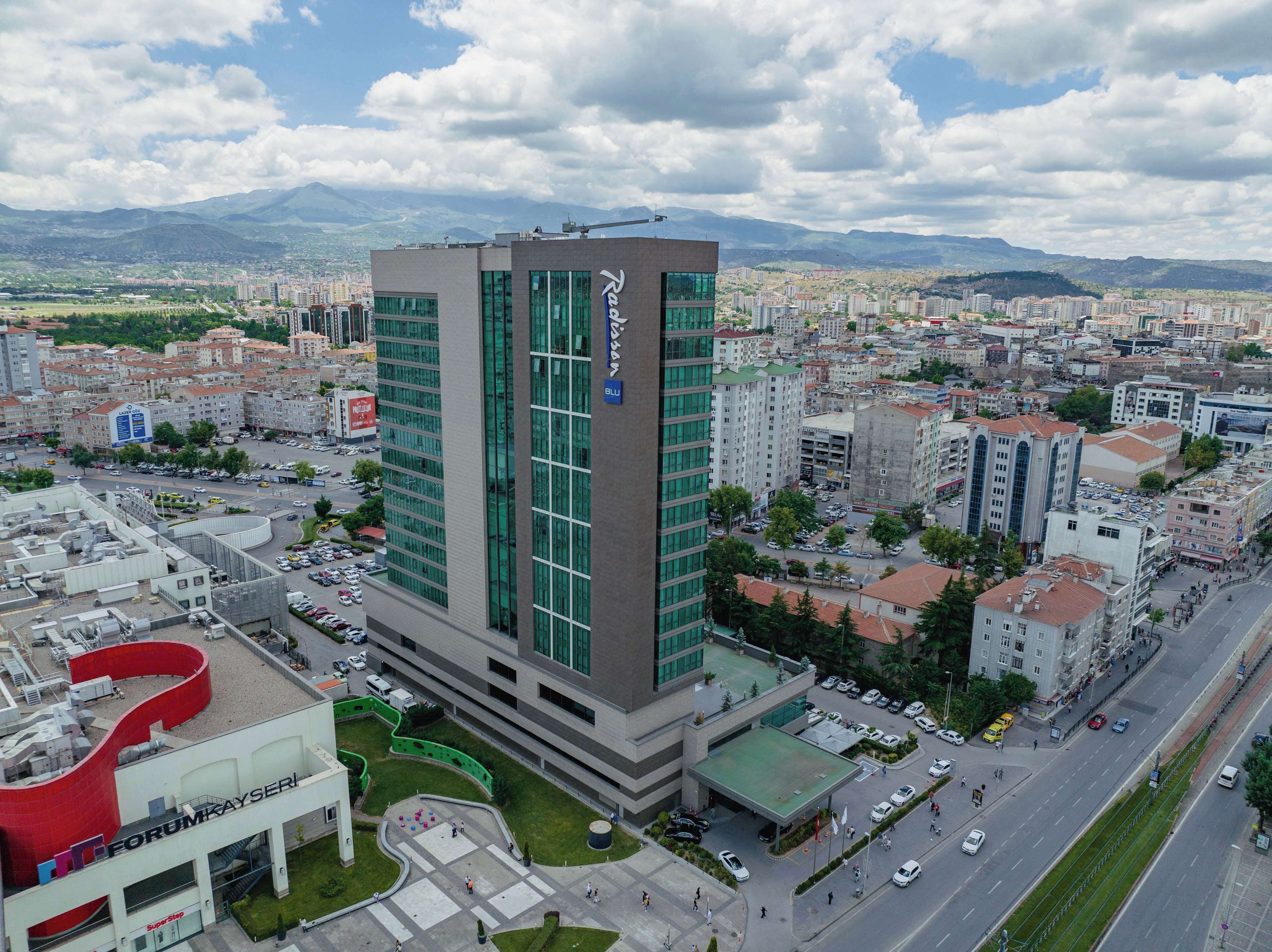 Un hôtel moderne doté d’une façade en verre remarquable, situé au cœur d’une ville et entouré d’un paysage urbain avec des montagnes en arrière-plan.
