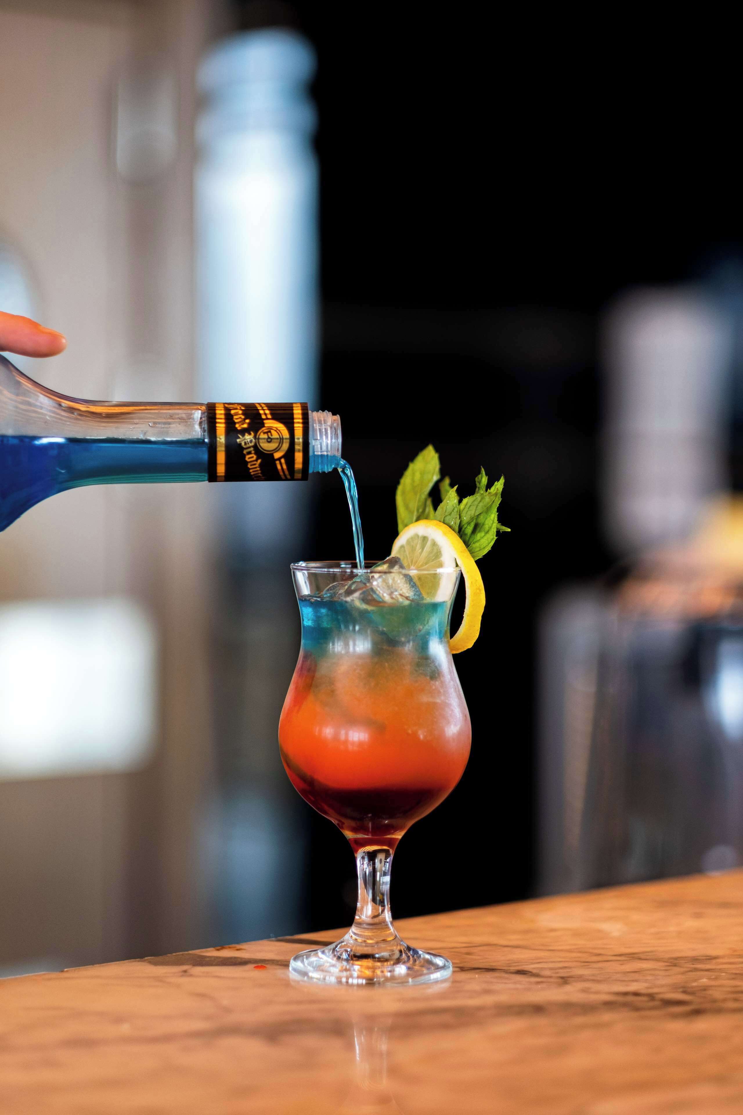 A cocktail being poured into a glass with a lemon slice and mint garnish.