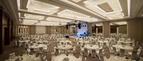 Un salón de banquetes con mesas redondas dispuestas para un evento formal, con un escenario y una gran araña.