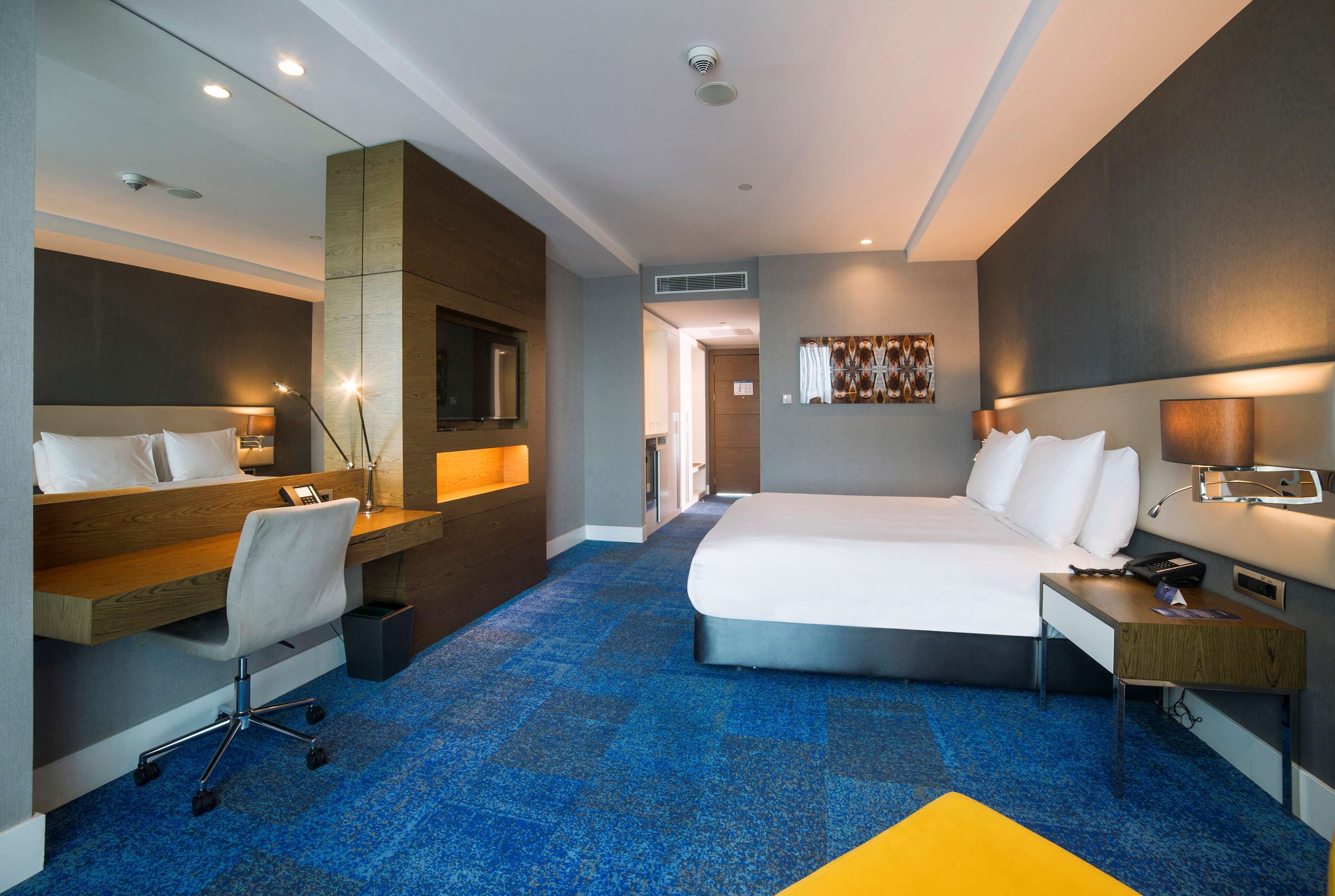 A modern hotel room with a large bed, a desk, a chair, and a small table.