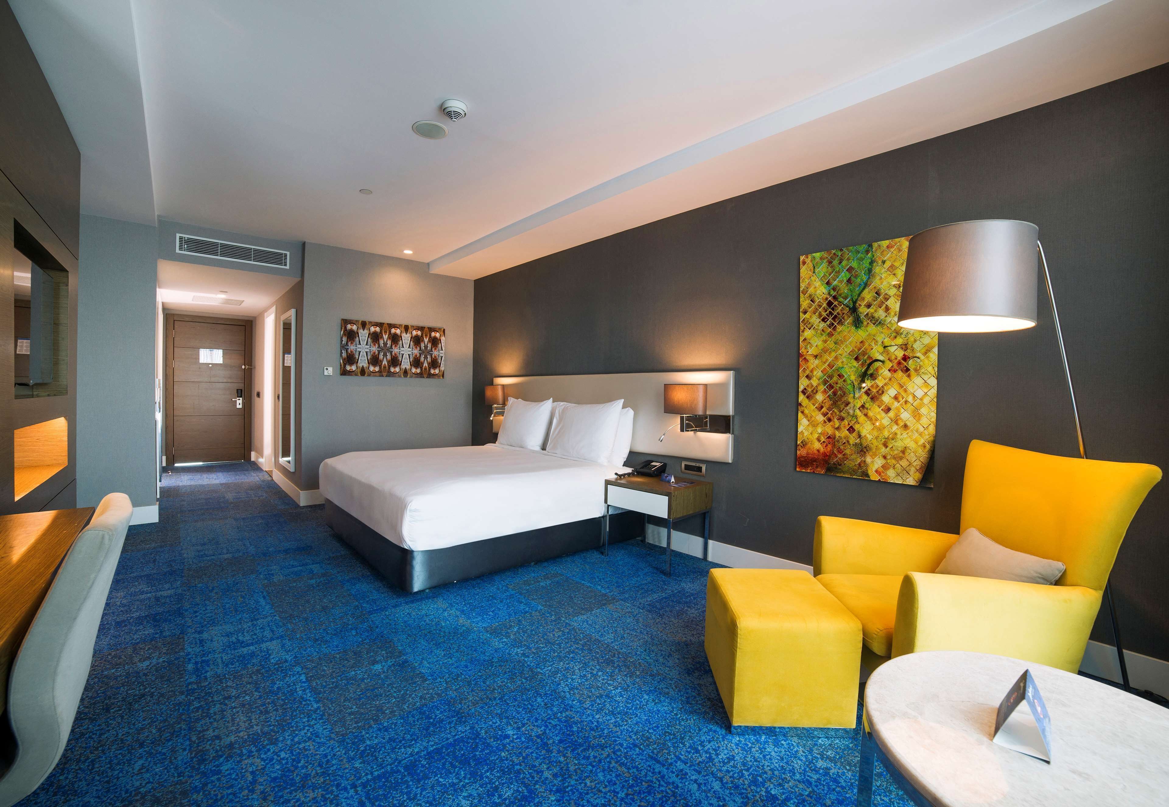 A hotel room with a large bed, a desk, a chair, a small table, and a lamp.