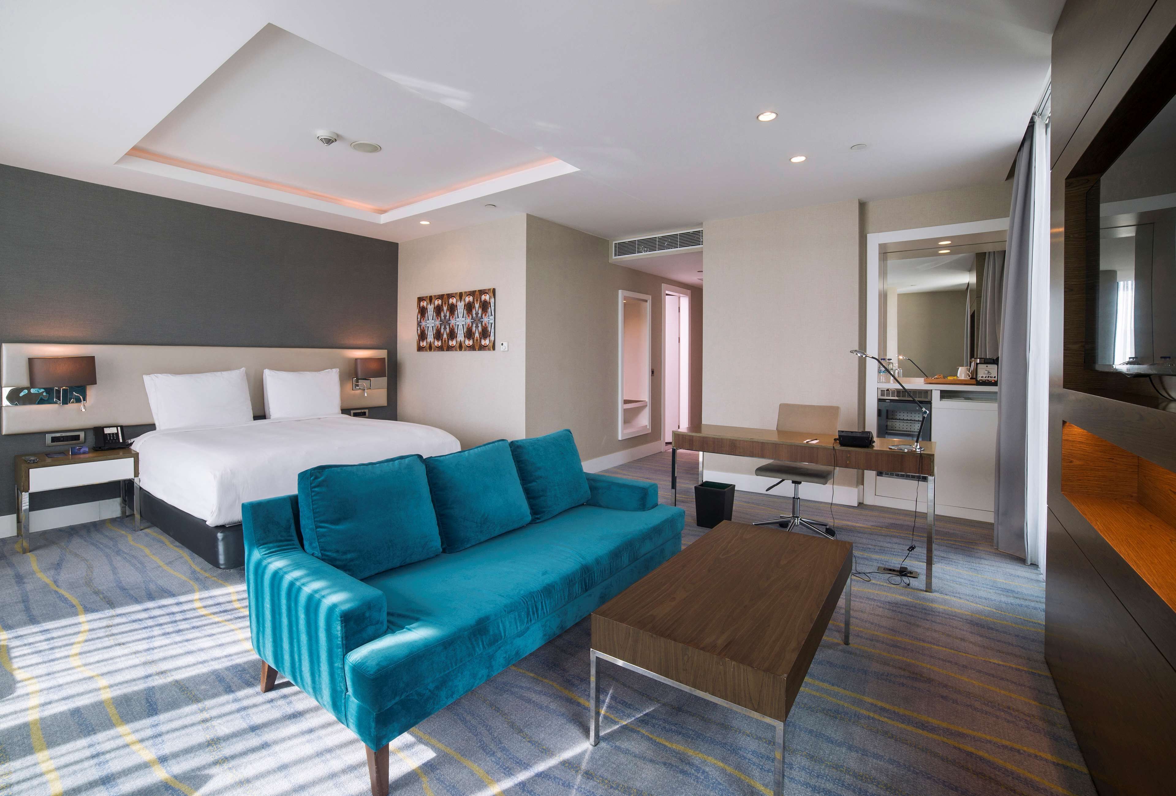 A modern hotel room with a large bed, a teal sofa, a wooden coffee table, a desk, and a large mirror.