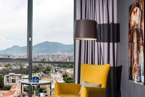 A room with a yellow armchair, a black floor lamp, a cityscape view, and a large painting on the wall.