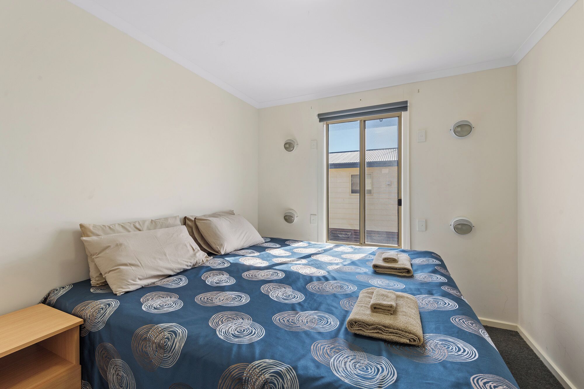 Un dormitorio con una cama, una mesita de noche de madera, un ventanal con vistas y una puerta.