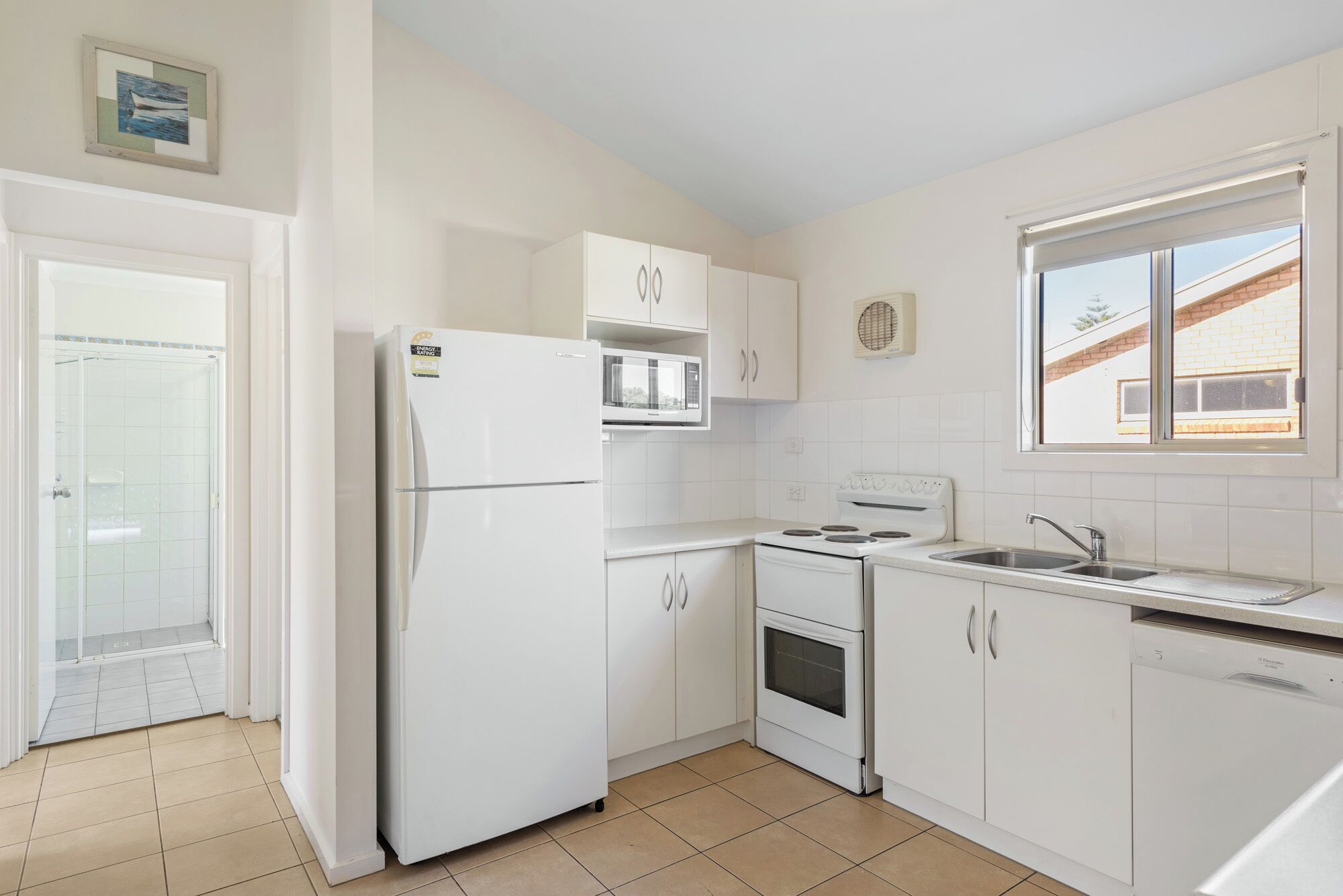 A small, white kitchen with a refrigerator, microwave, oven, and sink. There is a window with a view of a brick building outside.