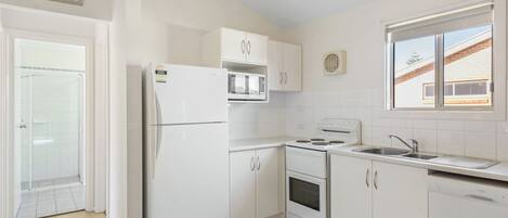 A small, white kitchen with a refrigerator, microwave, oven, and sink. There is a window with a view of a brick building outside.