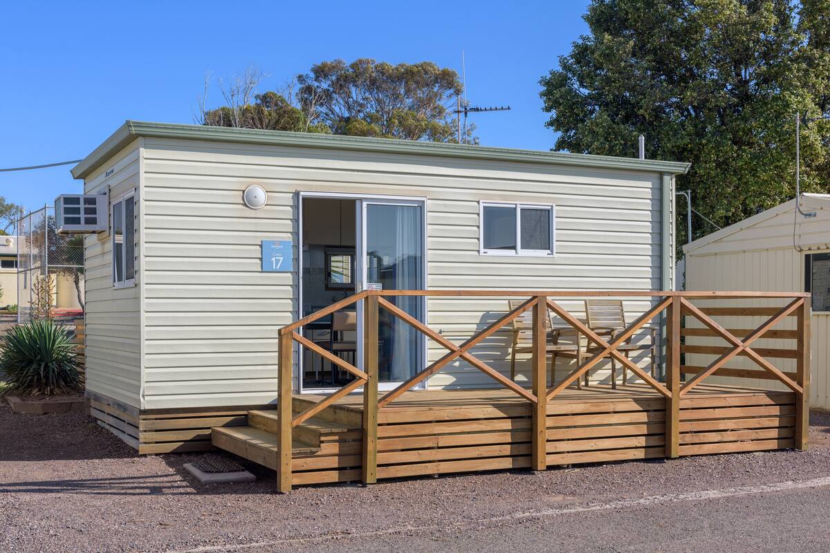 A small, single-story mobile home with a wooden deck and a door leading to a covered porch.