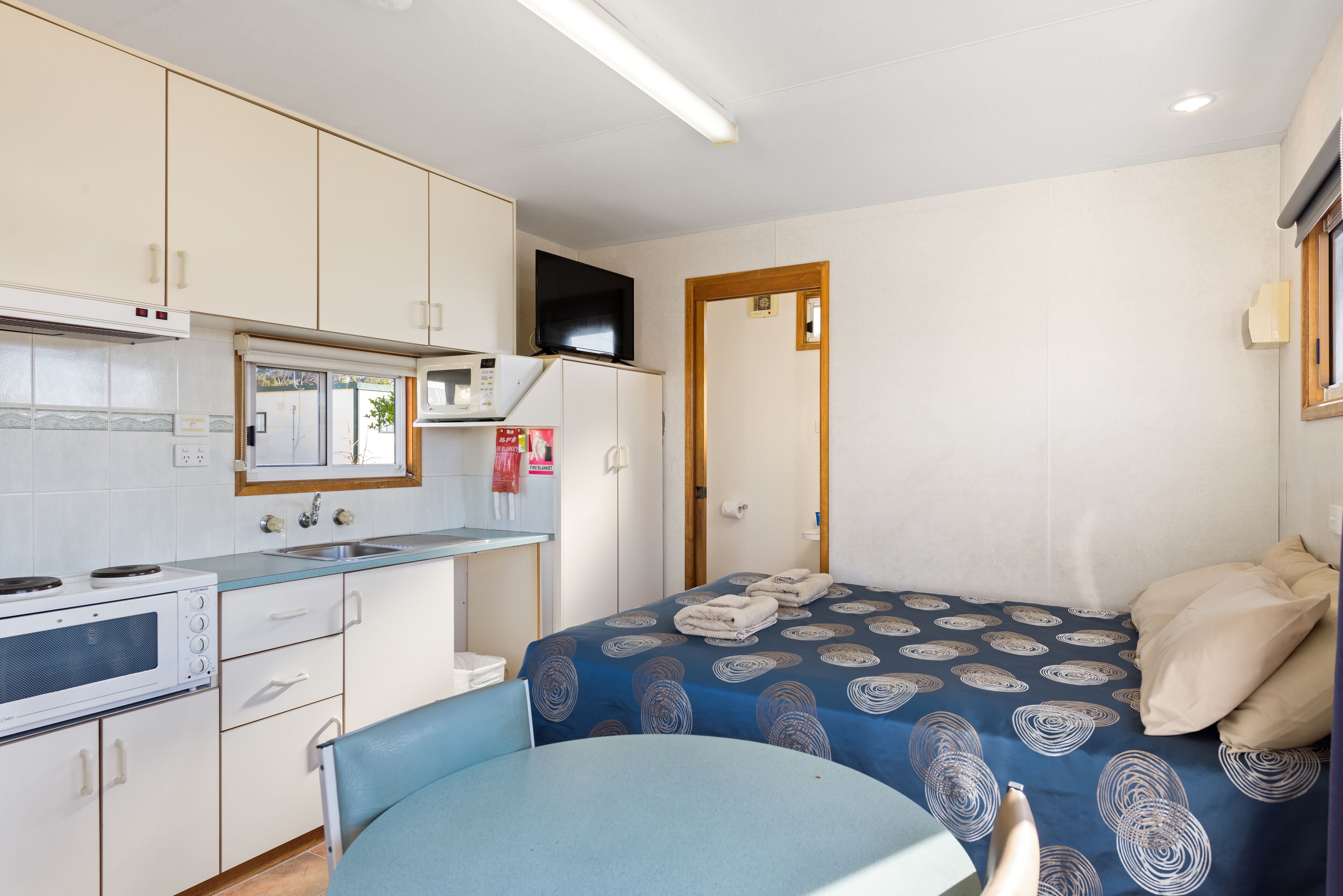 A compact hotel room with a kitchenette, a bed with a patterned bedspread, a small table, and a chair.