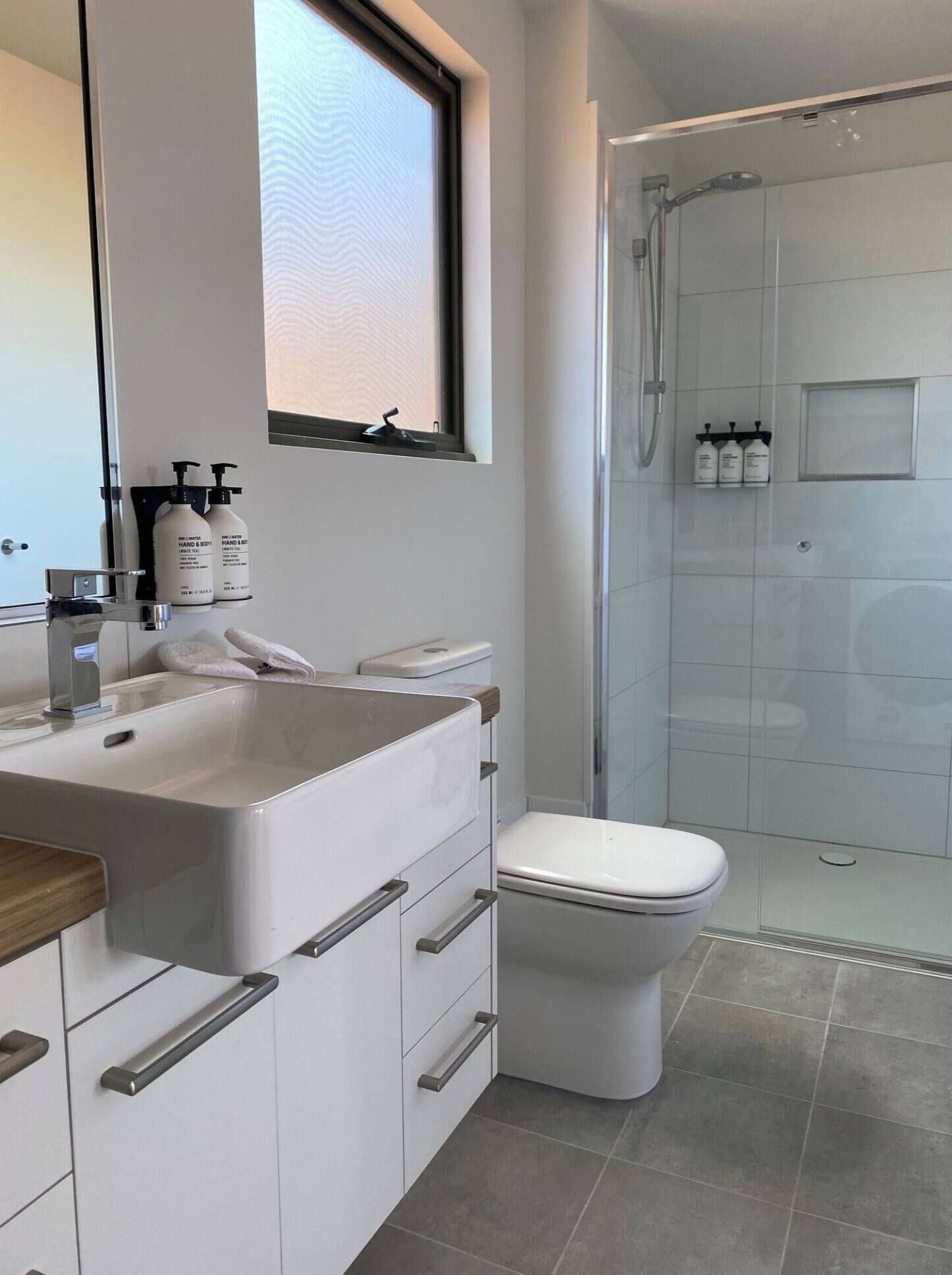 A modern bathroom with a white sink, toilet, and shower. There are hand and body soap dispensers on the sink and a window above it.