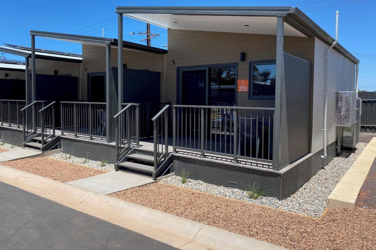 A modern building with a covered outdoor area, a sign indicating room number 203, and a small garden area with gravel and plants.
