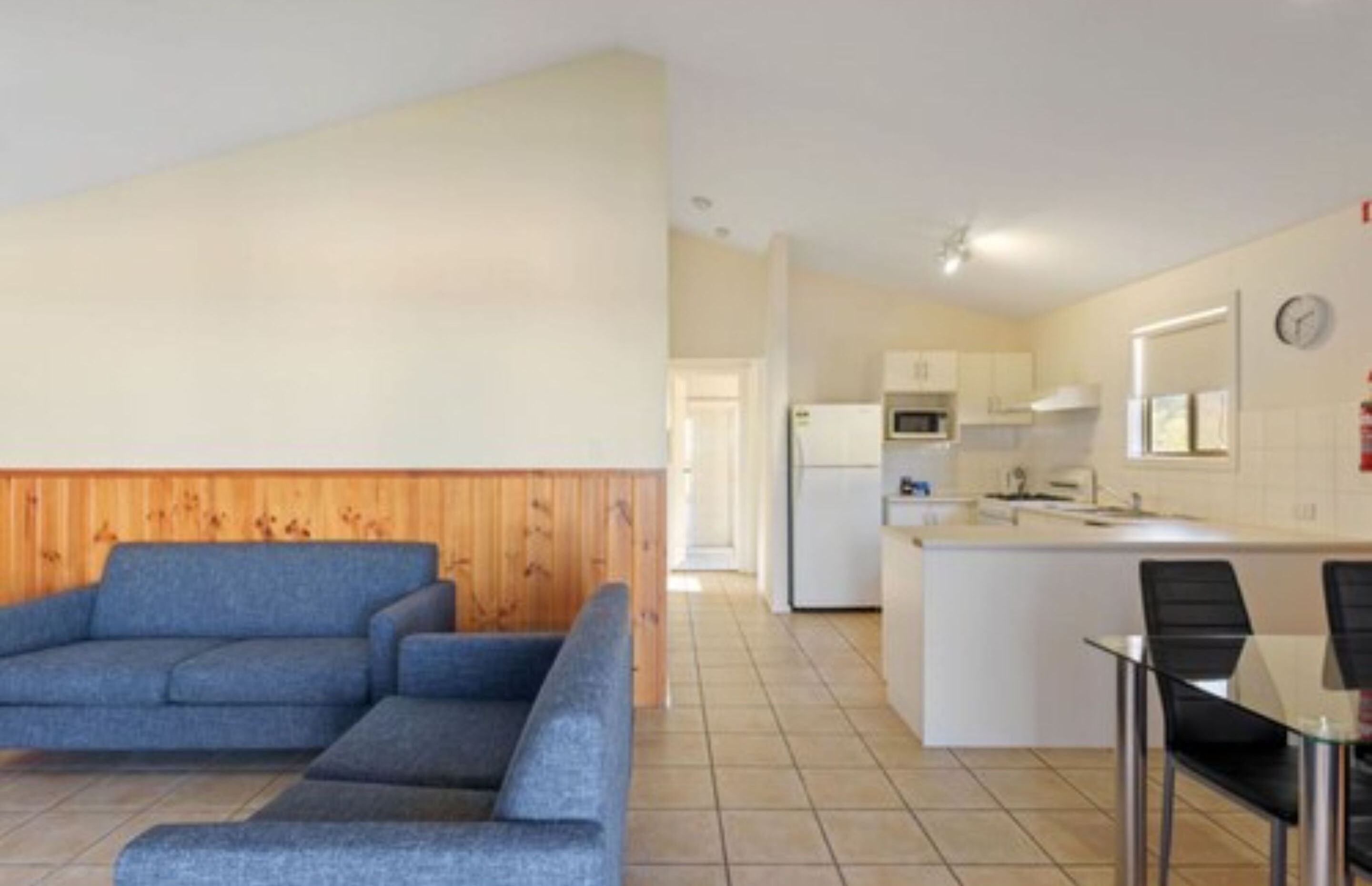 Un apartamento moderno con sala de estar que presenta un sofá azul, una pequeña cocina con electrodomésticos blancos y un comedor con mesa de vidrio y sillas negras.