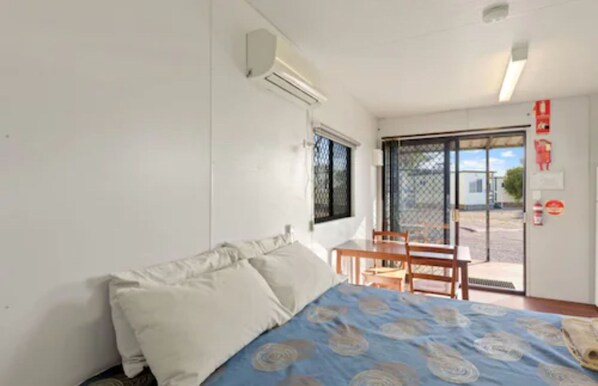 A room with a bed, a small table, and a chair. There is a window with a metal screen, and a door leading outside.