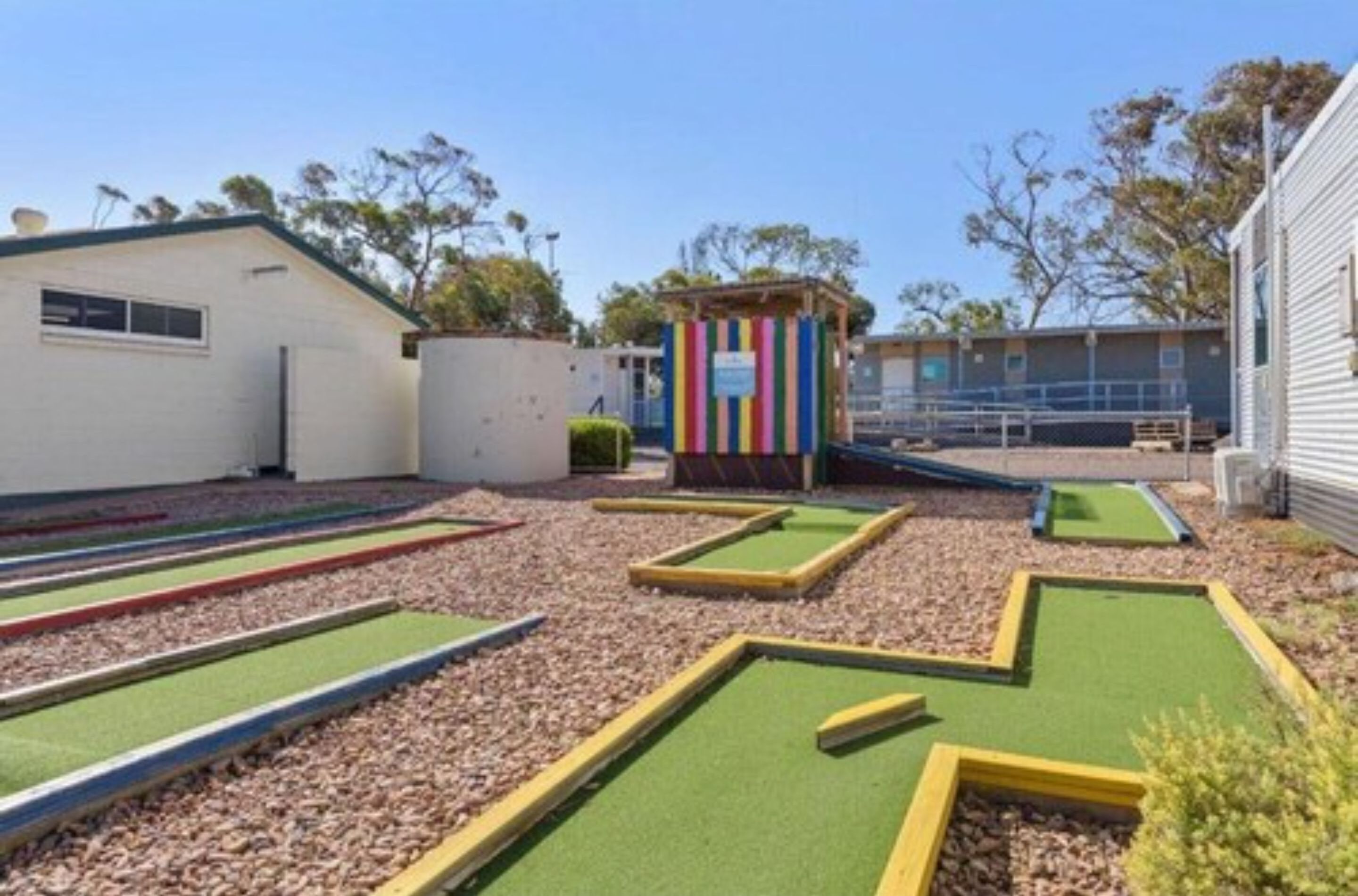 En minigolfbana med flera hål, en liten byggnad med en färgglad, randig fasad och en klar himmel.