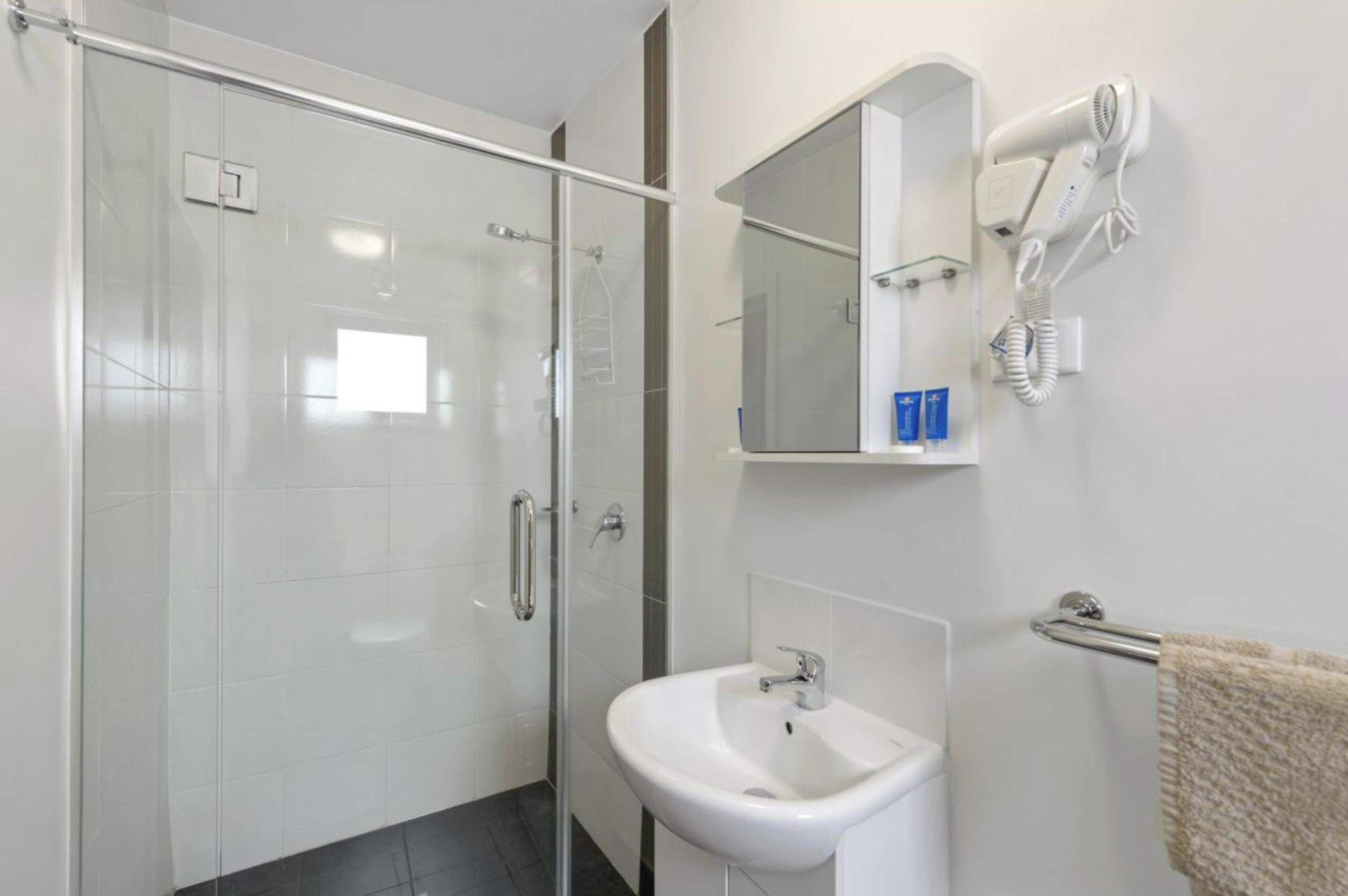 A modern bathroom with a glass shower enclosure, a white sink, a towel rack, and a hairdryer mounted on the wall.