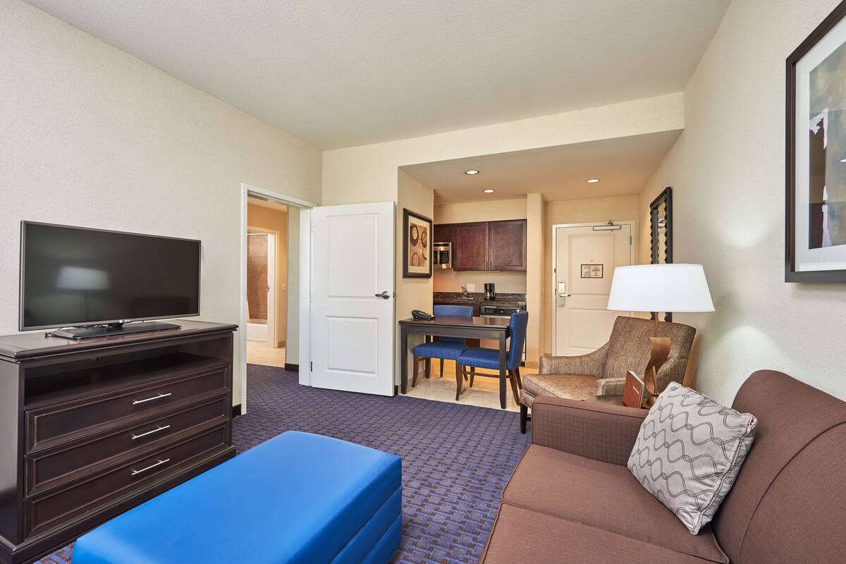 Habitación de hotel con televisor de pantalla plana, sofá, un reposapiés azul, zona de comedor con mesa y sillas, y área de cocina con armarios y microondas.