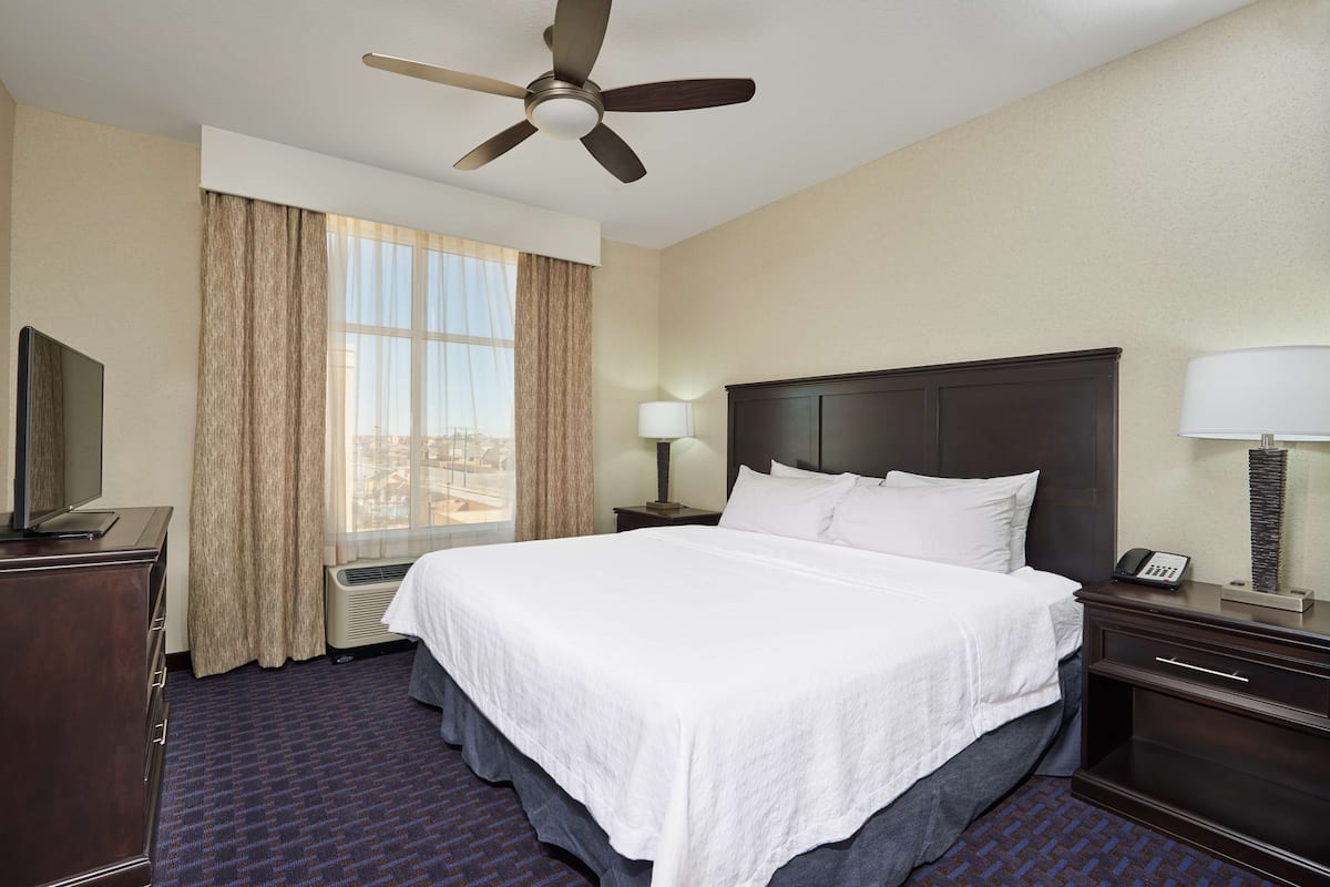 Habitación de hotel con una cama grande, un ventilador de techo, una ventana con cortinas, un escritorio con lámpara y un teléfono.