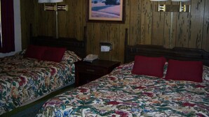 Quarto de hotel com duas camas, cada uma com roupa de cama floral e almofadas vermelhas. Há duas luminárias fixadas na parede e um quadro emoldurado sobre as camas.