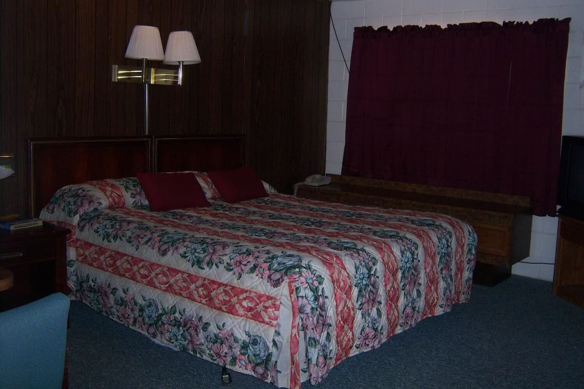 A hotel room with a wooden headboard, a floral bedspread, a pair of lamps, and a red curtain.