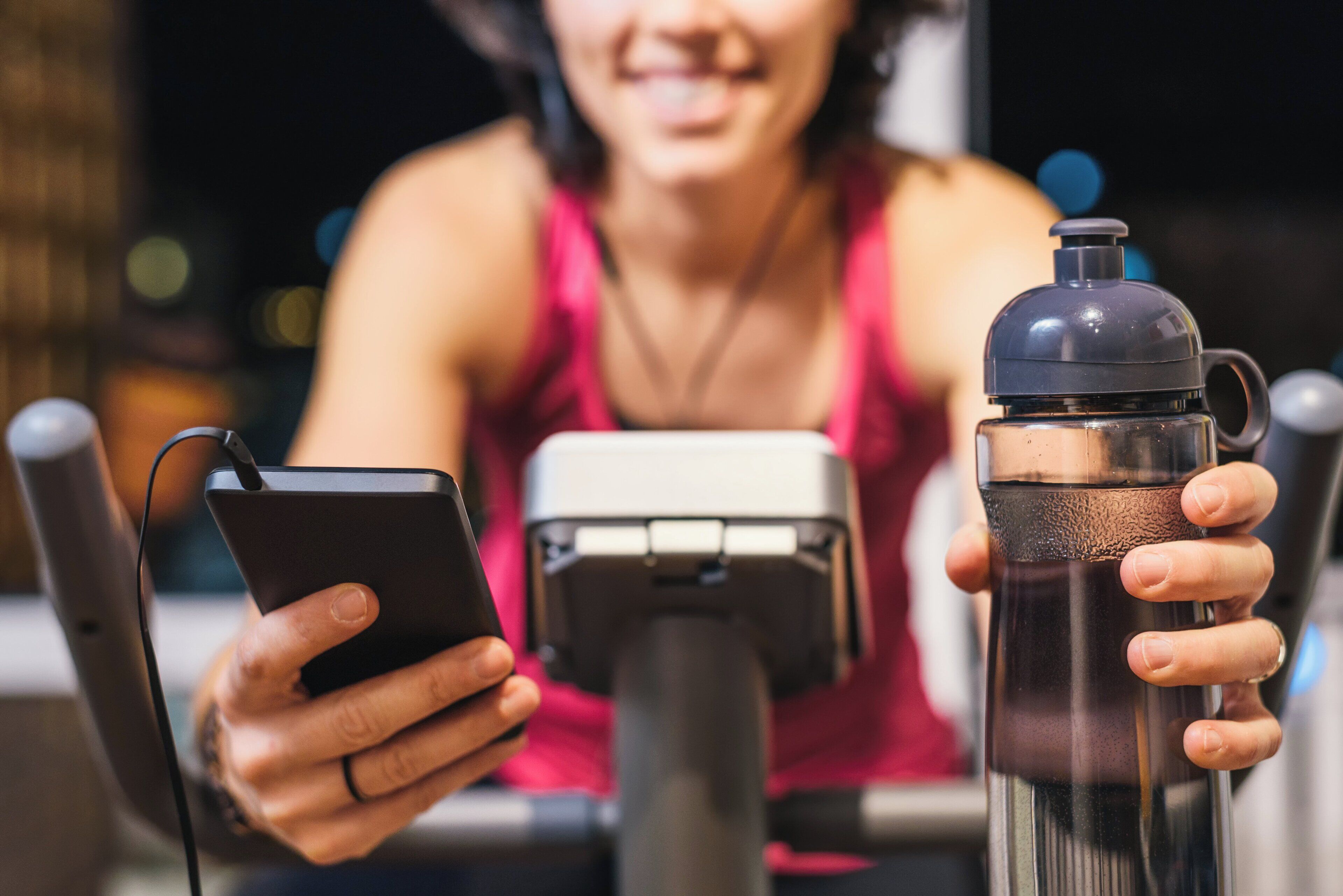 A person holding a smartphone and a water bottle, with a stationary bike in the background.