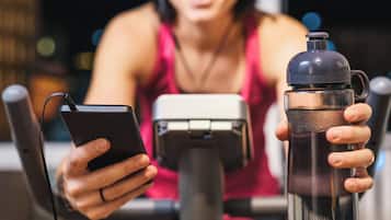 A person holding a smartphone and a water bottle, with a stationary bike in the background.