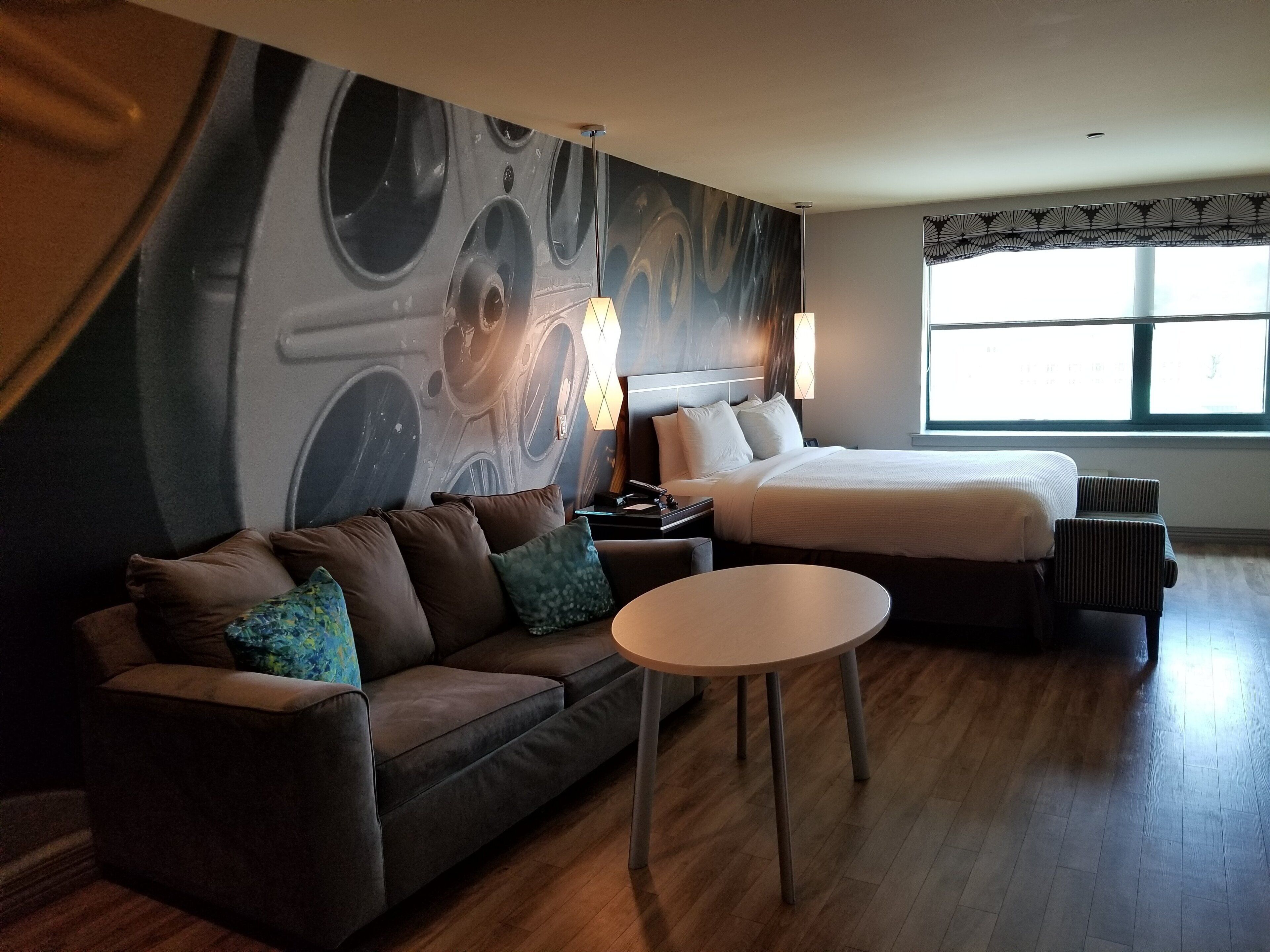 A modern hotel room with a large bed, a sofa, a small table, and a wall mural.