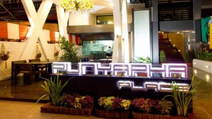 Un moderno vestíbulo con una recepción identificada con el rótulo “PLAYAH PLACE”, que cuenta con una araña, plantas en macetas y una escalera.