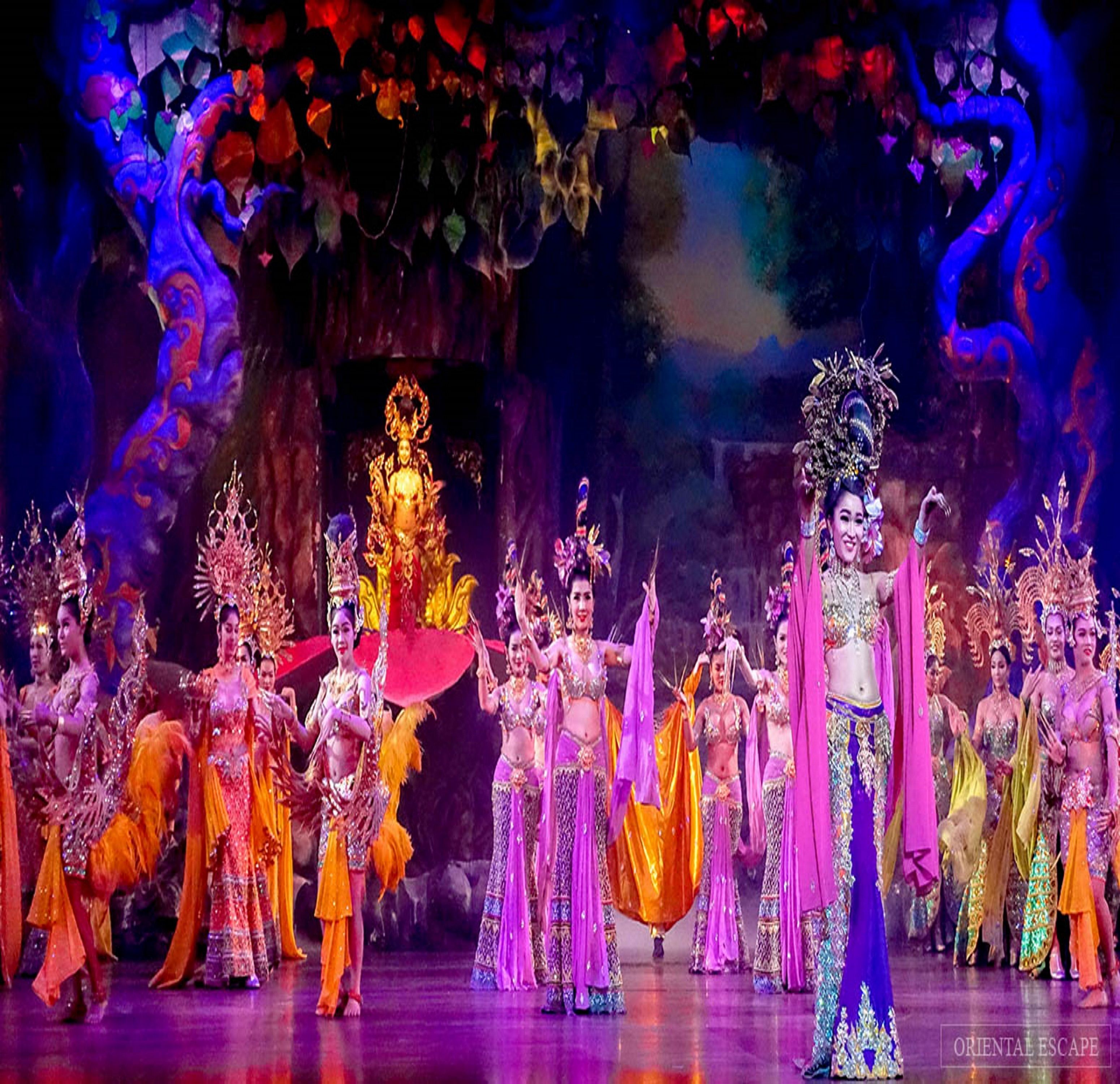 A group of performers in traditional costumes on a stage with a backdrop of colorful decorations and a central figure in elaborate attire.