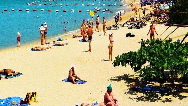 Una playa con gente tomando el sol y nadando, palmeras y un cielo azul despejado.