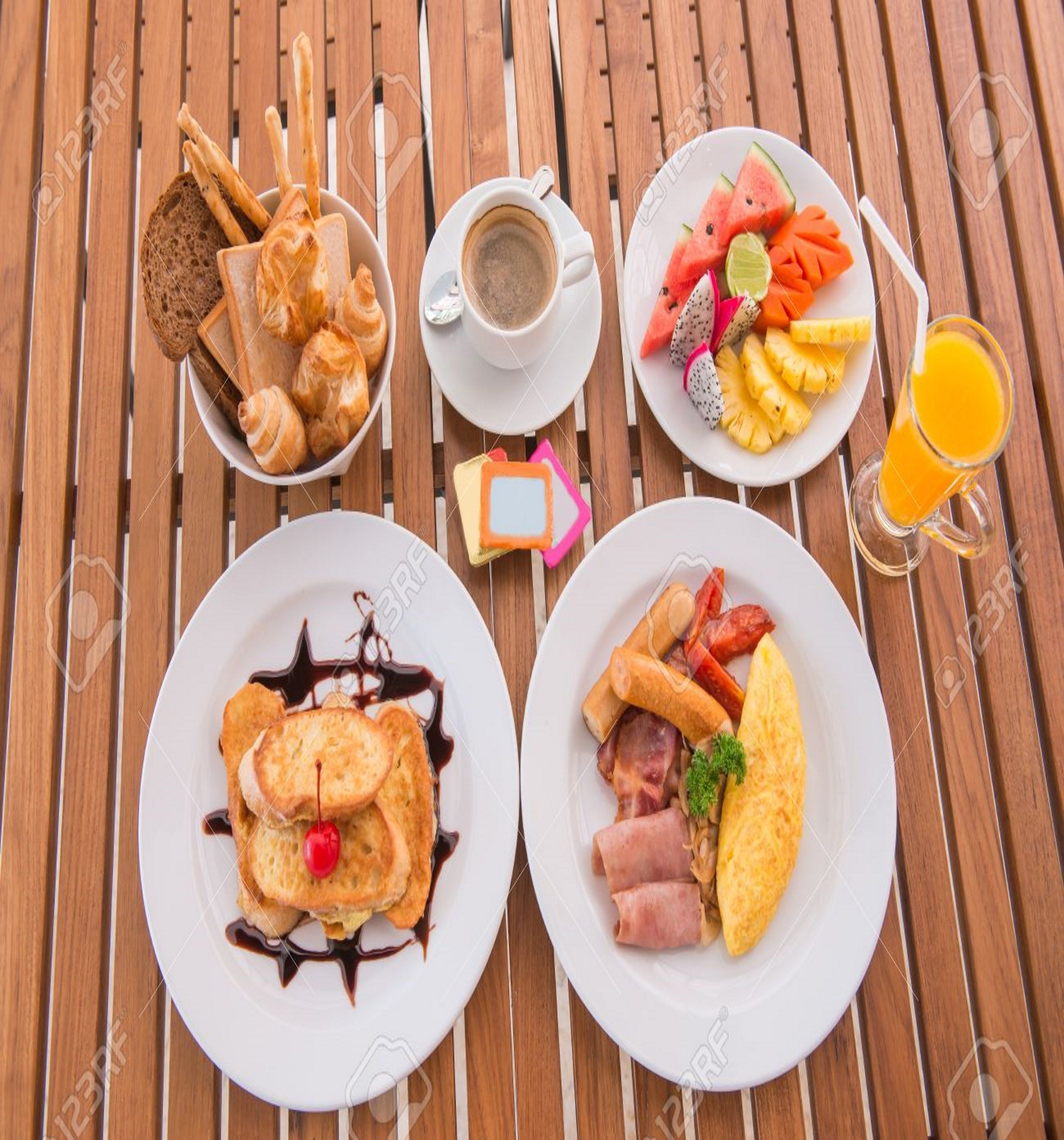 A breakfast spread with croissants, coffee, fruit, eggs, and pancakes on a wooden table.