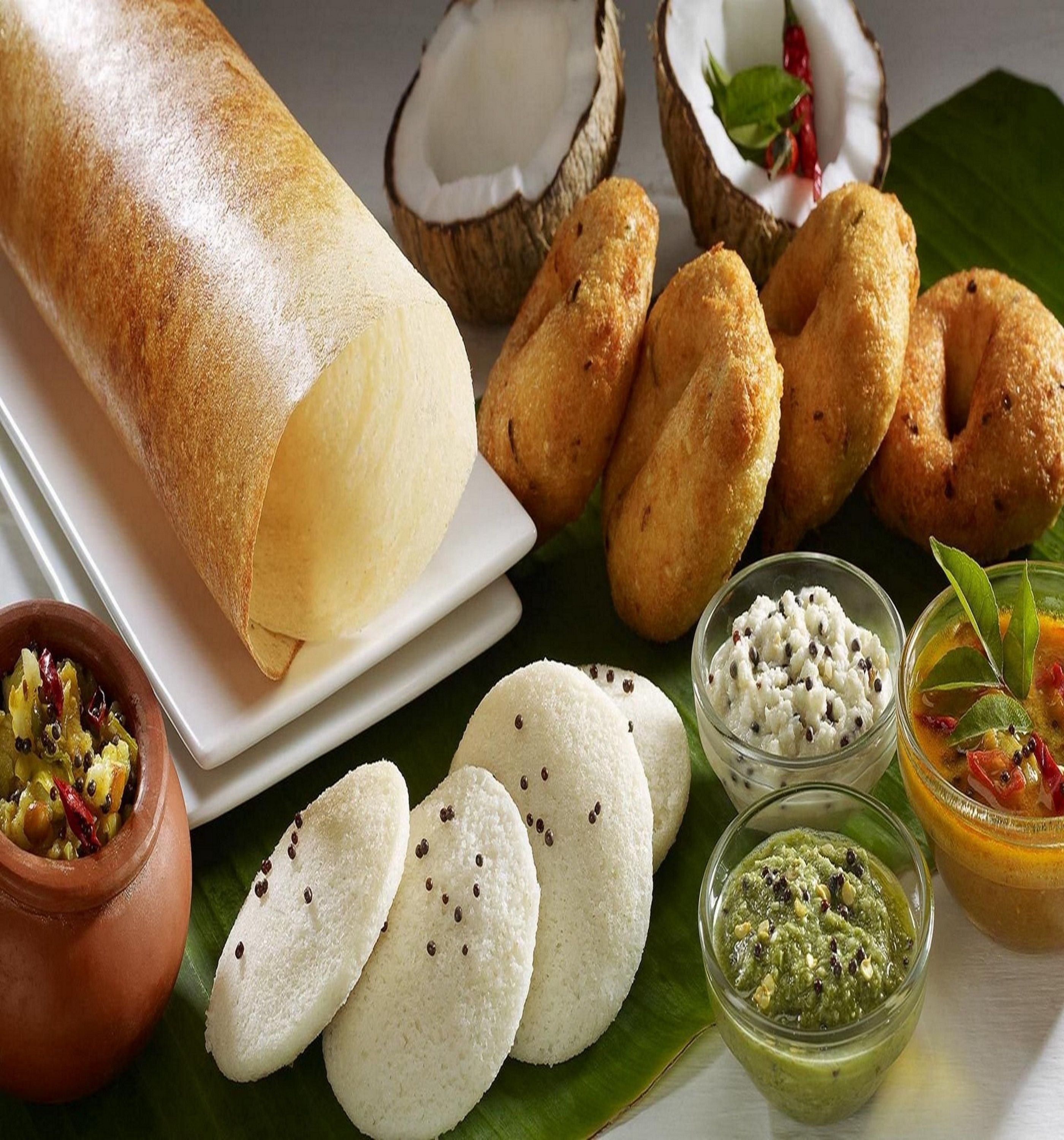 A variety of South Indian snacks and dishes, including a large dosa, coconut halves, and several types of idlis and vadas.