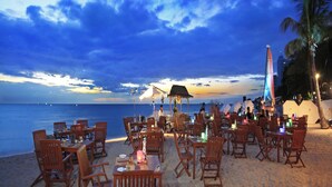 Un área de comedor al aire libre con mesas y sillas dispuestas sobre la arena, con vista al océano.