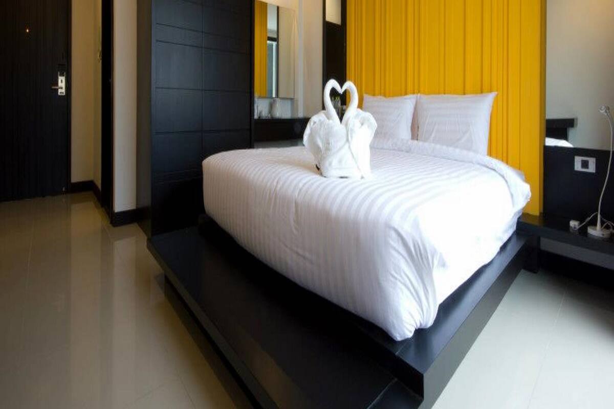 Une chambre d’hôtel moderne avec un grand lit, une armoire noire, un mur d’accent jaune et une serviette blanche disposée en forme de cœur.