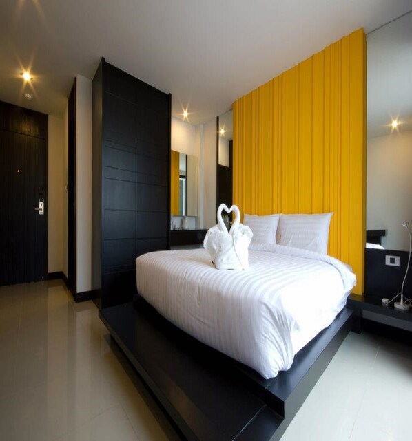 A modern hotel room with a large bed, a black wardrobe, a yellow accent wall, and a white heart-shaped towel arrangement.