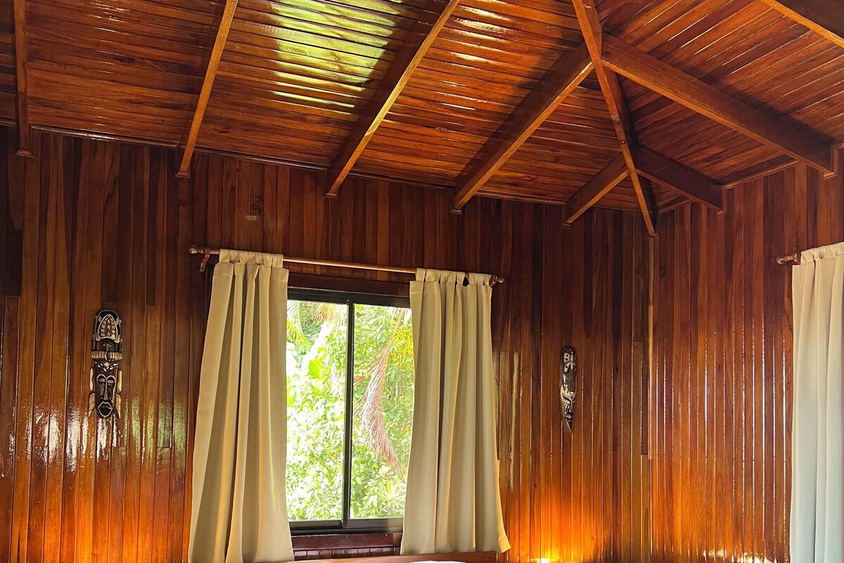 A wooden cabin room with a bed, curtains, a window, and a ceiling fan.