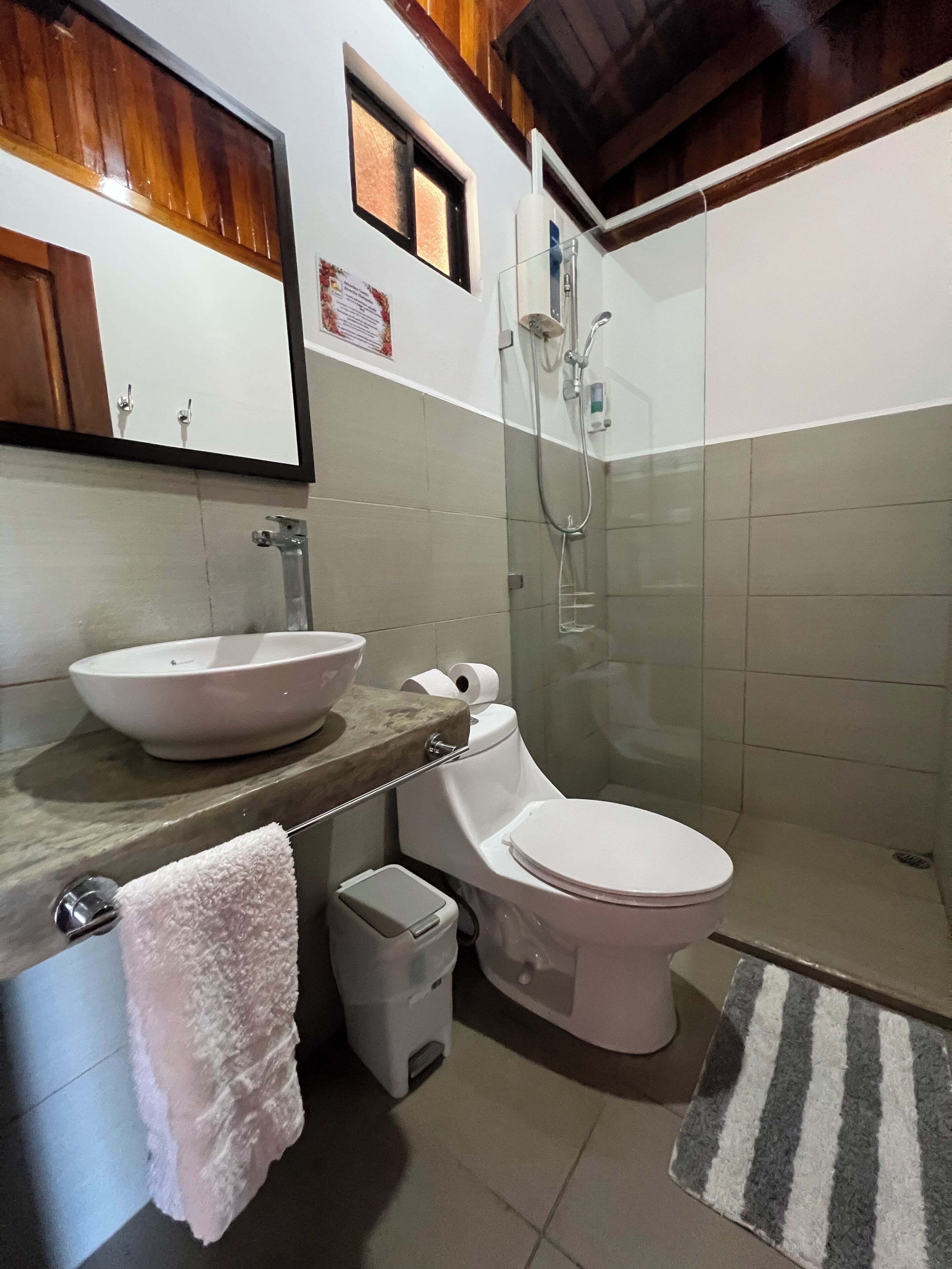 Casa de banho com lavatório branco, sanita e chuveiro. Há um porta-toalhas com toalha branca, um contentor de lixo e um tapete riscado no chão.