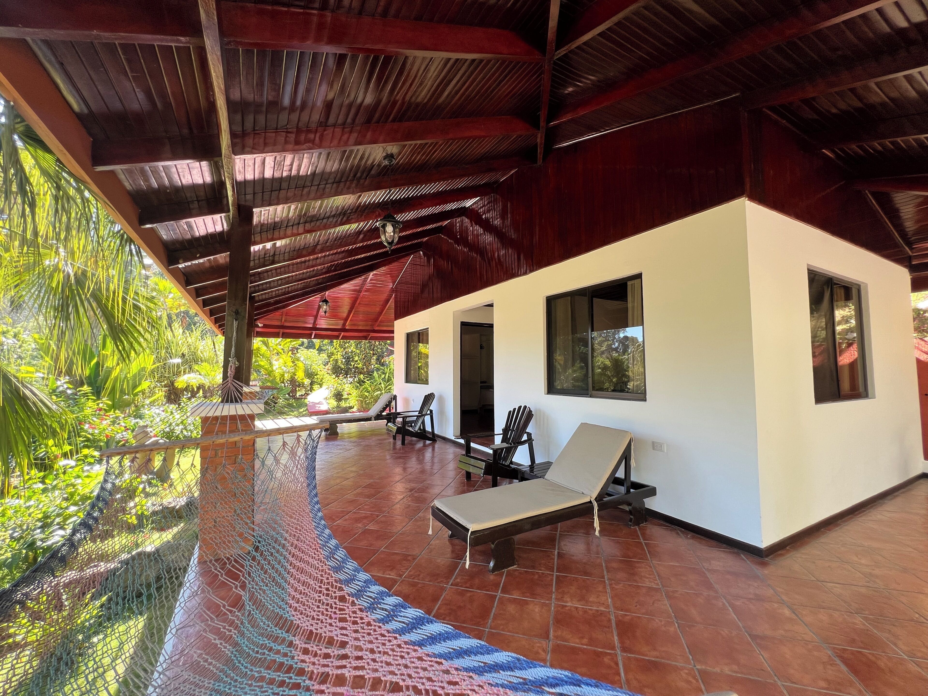 A covered outdoor area with a pool, lounge chairs, and a white building with windows.