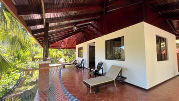 A covered outdoor area with a pool, lounge chairs, and a white building with windows.