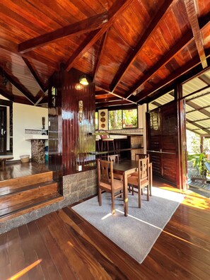 A spacious room with a high ceiling, wooden beams, a dining area with chairs and a table, and a view of the outdoors through large windows. - Hotel El Jardín (Cóbano)
